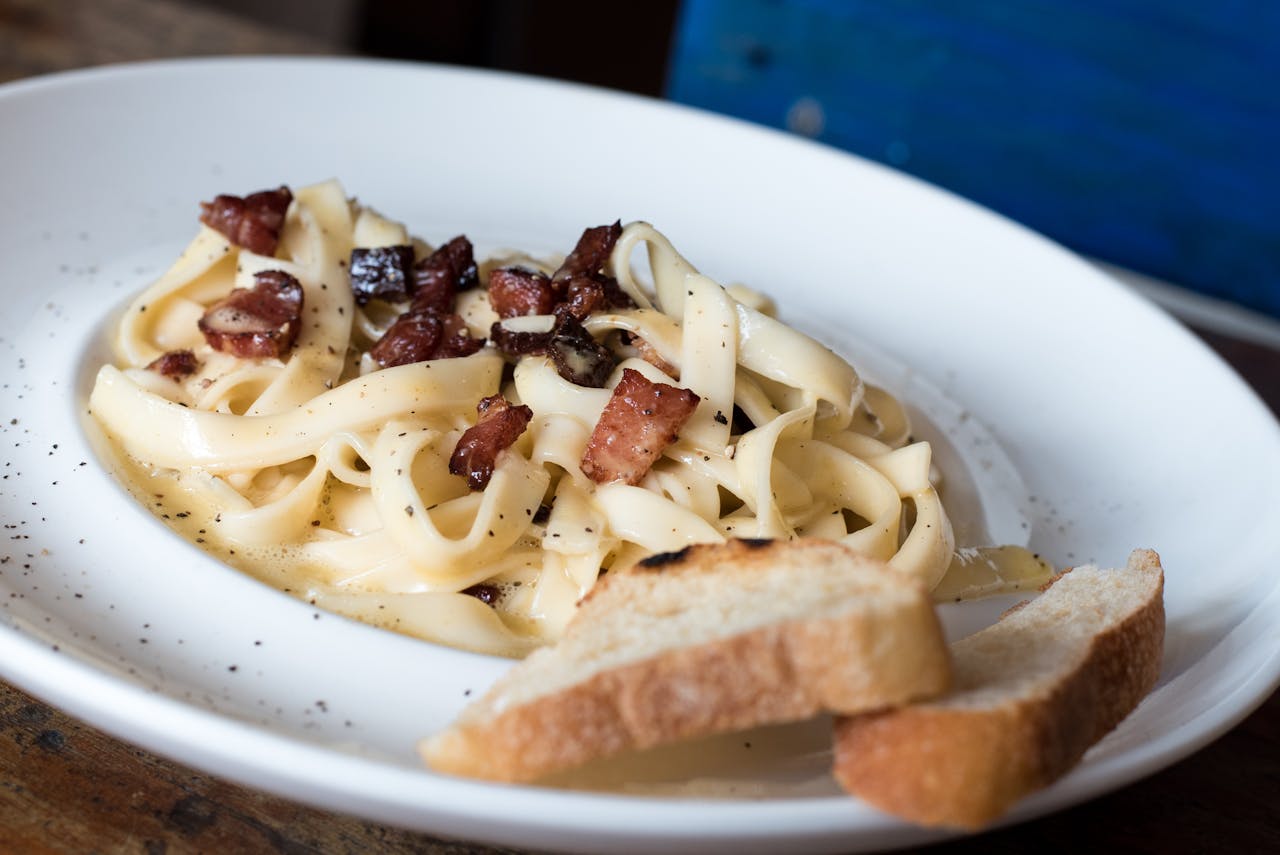 Close-up of creamy carbonara pasta topped with bacon, served with fresh bread in a restaurant.